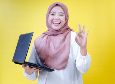 Tesettürlü neşeli Asyalı bir kadın dizüstü bilgisayarını tutuyor ve sarı arka plana karşı iyi bir el hareketi gösteriyor.