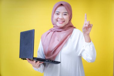 tesettürlü gülümseyen Asyalı bir kadın dizüstü bilgisayarını tutuyor, gülümsüyor ve sarı arka plana doğru işaret ediyor..