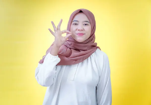 Başörtülü gülümseyen Asyalı bir kadın parlak sarı bir arka planda el işareti gösteriyor.