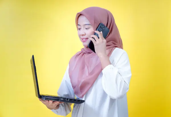 Sarı arka planda dizüstü bilgisayarını tutan tesettürlü genç Asyalı bir kadın telefonda konuşuyor..