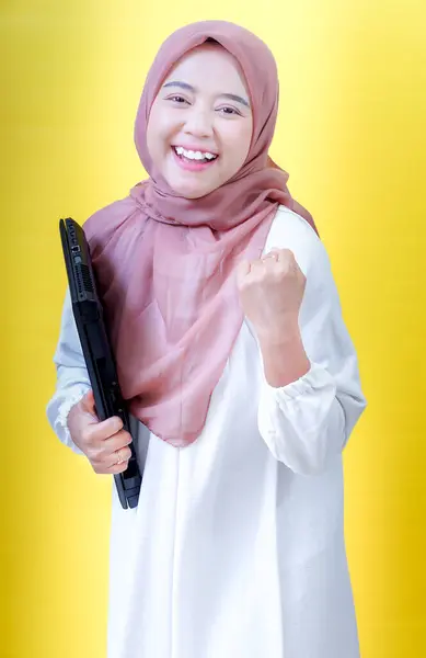 Beyaz elbiseli ve tesettürlü neşeli Asyalı bir kadın dizüstü bilgisayar tutuyor, sarı bir arka plana karşı başarıyı kutluyor..