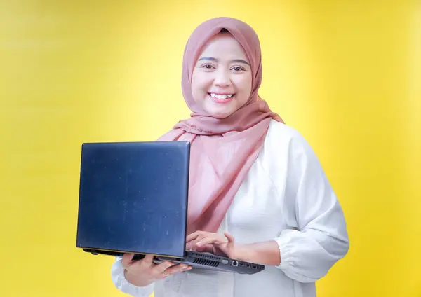 Tesettürlü gülümseyen Asyalı bir kadın dizüstü bilgisayar tutuyor, parlak sarı bir arkaplanda..