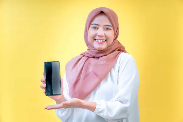 Tesettürlü gülümseyen Asyalı bir kadın akıllı telefonunu parlak sarı bir arka planda sergiliyor.