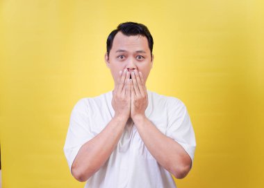 Şaşırmış Asyalı bir adam parlak sarı bir arka planda ağzını kapatıyor.