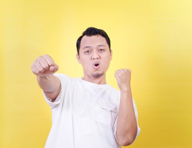 Asyalı bir adam, canlı sarı bir arka plana karşı yumruklarıyla heyecan gösteriyor.