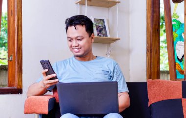 Kapalı bir ortamda akıllı telefon ve laptop kullanan gülümseyen Asyalı bir adam.