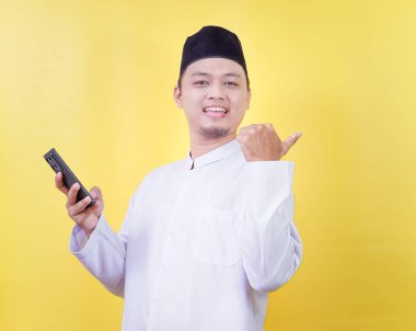 A smiling Asian Muslim man in a white prayer garment gives a thumbs up to the rear while holding smartphone clipart