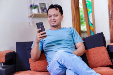 Kanepede rahat bir şekilde oturan Asyalı bir adam, akıllı telefonuyla meşgul ve gülümsüyor..