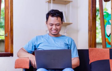 Genç Asyalı bir adam rahat bir odada dizüstü bilgisayar kullanırken gülümsüyor..
