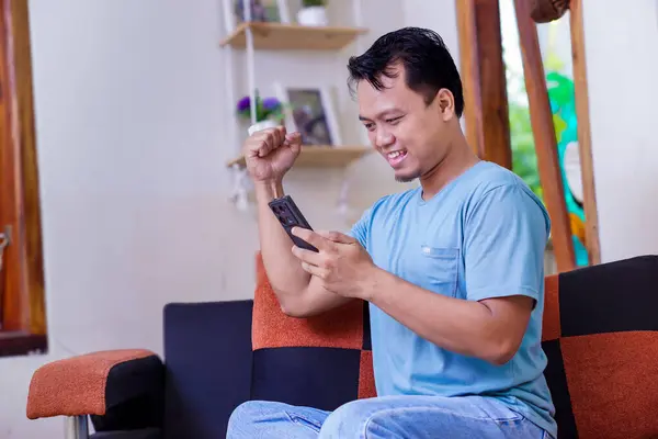 Asyalı bir adam evinde akıllı telefonunu kullanarak zaferi kutluyor, coşku ve neşe gösteriyor.