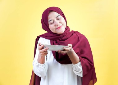 An asian muslim woman in a hijab joyfully holds a plate, expressing happiness against a yellow background. clipart