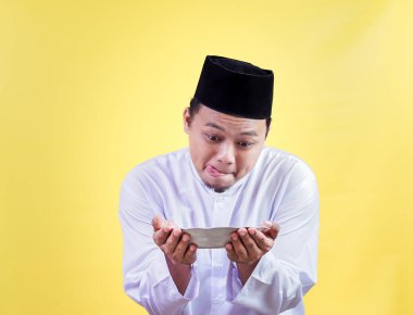 Geleneksel kıyafetli Asyalı Müslüman bir adam, parlak sarı bir arka plana karşı bir tabağa aç bir şekilde bakar.