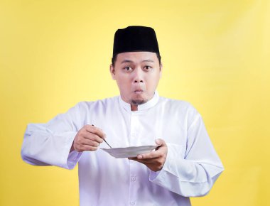 An asian muslim man in traditional attire eagerly enjoys a meal, showcasing a moment of delight clipart