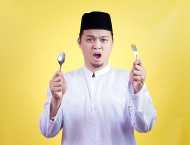 A surprised asian muslim man in traditional attire holds a spoon and fork against a yellow background clipart