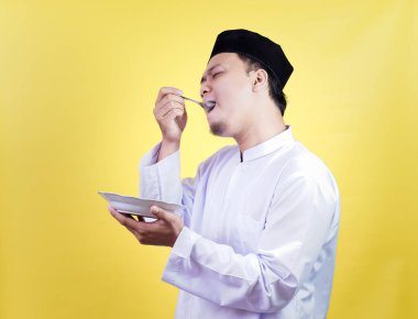 Geleneksel giyinmiş Asyalı bir Müslüman, sarı arka planda tatmin olmuş bir ifadeyle lezzetli yemeklerden hoşlanır.