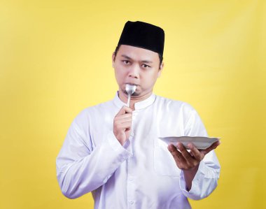 Geleneksel giysili Asyalı Müslüman bir adam elinde tabak tutarken kaşığı ısırıyor.