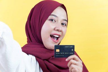 Mutlu Asyalı tesettürlü Müslüman bir kadın kredi kartını güçlü bir şekilde sarı arka planda tutarken selfie çekiyor..