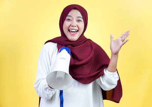 Neşeli Asyalı Müslüman bir kadın tesettüre sarılı bir megafonla parlak sarı bir zemin tutuyor.