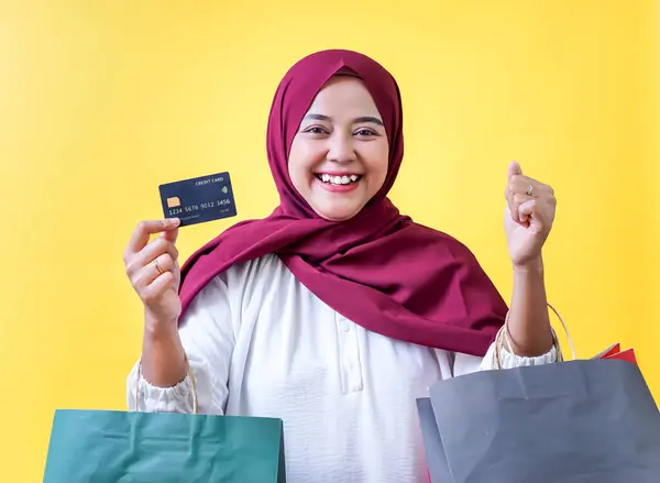 Tesettürlü Asyalı Müslüman bir kadın alışveriş torbaları ve sarı arka planda bir kredi kartı tutuyor..