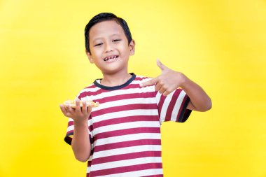 A cheerful asian boy in a striped shirt holds a slice of pizza and gives a thumbs up against a bright yellow background. international foods day concept clipart