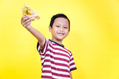 A cheerful asian boy holds up a slice of pizza against a bright yellow background, spreading joy. international foods day concept clipart