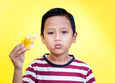 Asyalı genç bir çocuk parlak sarı bir arka planda lezzetli bir dilim pizzanın tadını çıkarıyor. Uluslararası Gıda Günü konsepti