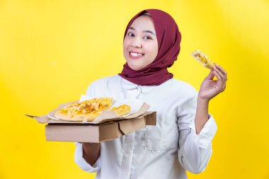 Tesettürlü neşeli bir Asyalı kadın bir kutuda pizzayı parlak sarı bir arka planda tutuyor.