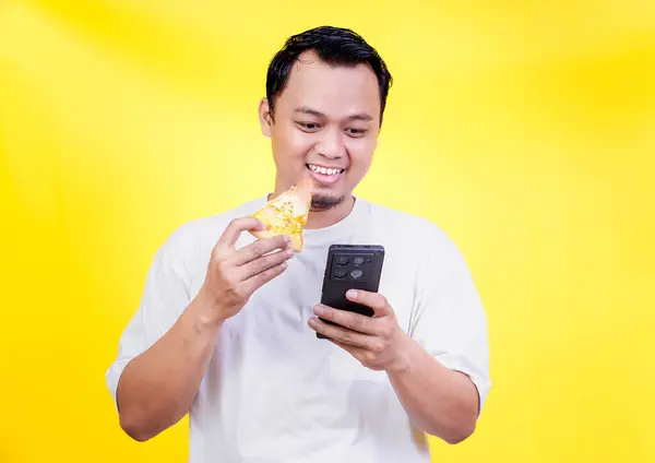 Gülümseyen Asyalı adam telefonuna parlak sarı bir arka planda bakarken pizzadan zevk alır. Uluslararası Gıda Günü konsepti