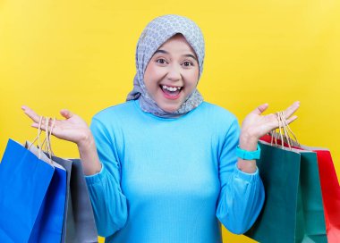 Alışveriş torbalarıyla tesettürlü neşeli Asyalı bir kadın, parlak sarı bir arka planda eğlenceli bir alışveriş deneyimi yaşıyor..