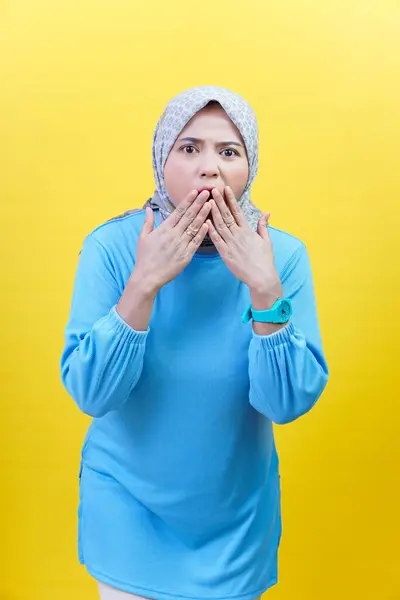 Tesettürlü ve mavi elbiseli Asyalı bir kadın şok olmuş görünüyor, iki eliyle sarı arka planda ağzını kapatıyor.