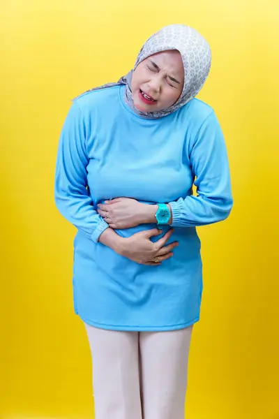 Sarışın ve mavi elbiseli Asyalı bir kadın, sarı arka planda rahatsız olduğunu belirterek, karnını acı içinde tutuyor..