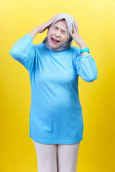 Tesettürlü ve mavi elbiseli Asyalı bir kadın hayal kırıklığını ifade eder. İki eliyle sarı arka planda başını tutar..