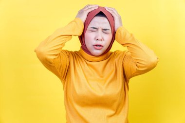 Kahverengi tesettürlü ve sarı kazaklı stresli bir Asyalı kadın başını kapalı gözlerle tutuyor..