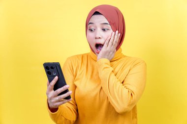 Şok olmuş, tesettürlü Asyalı bir kadın, akıllı telefonuyla sarı arka planda şaşırtıcı bir şeye tepki veriyor..