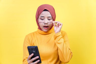 Yüzünde endişeli bir ifadeyle telefonuna bakarken tesettürlü Asyalı bir kadın gözlüğünü ısırıyor..