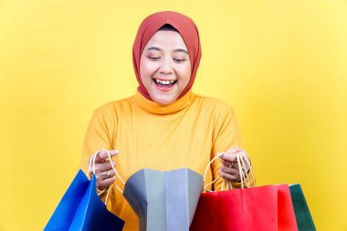 Tesettürlü neşeli Asyalı bir kadın parlak sarı bir arka planda renkli alışveriş torbaları tutuyor..