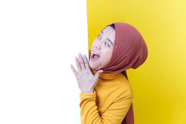 Tesettür ve hardal süveter giymiş neşeli Asyalı bir kadın boş beyaz bir duvara bakar ve şaşırdığını ifade eder..