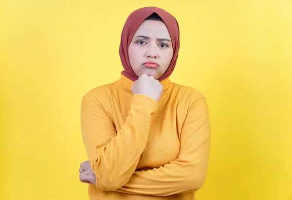 Kahverengi tesettürlü ve sarı kazaklı Asyalı bir kadın. Üzücü bir ifade sergiliyor. Sarı arka planda dalgın bir duruşu var..