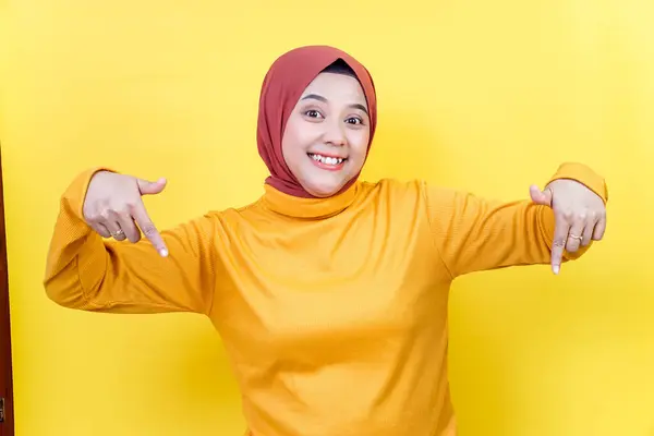 Kahverengi tesettürlü ve gözlüklü neşeli Asyalı bir kadın her iki elini de sarı arka plana doğru uzatıyor..