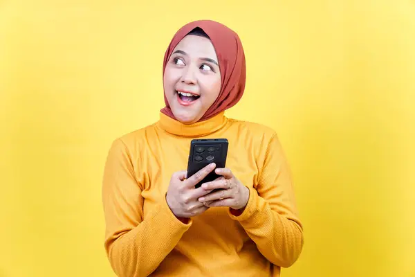 Tesettürlü neşeli bir Asyalı kadın, parlak sarı bir arka plana karşı akıllı telefonunu tutarken heyecanlı görünüyor..