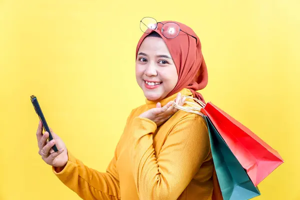 Tesettürlü neşeli bir Asyalı kadın, elinde alışveriş çantaları ve akıllı bir telefonla eğlenceli bir alışveriş deneyimi yaşıyor..