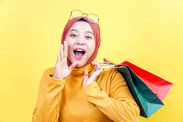 Tesettürlü neşeli bir Asyalı kadın elinde renkli çantalar tutarken heyecanla alışveriş yaptığını ilan ediyor..