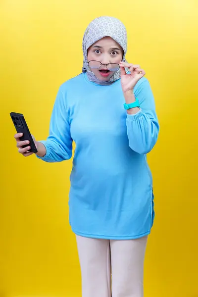 Mavi elbiseli ve tesettürlü şaşırmış bir Asyalı kadın şok içinde akıllı telefonuna bakarken gözlüklerini indiriyor..