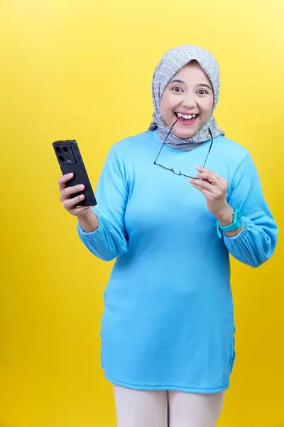 Mavi elbiseli ve tesettürlü neşeli Asyalı bir kadın telefonunu ve gözlüklerini tutuyor, heyecanla ve neşeyle tepki veriyor..