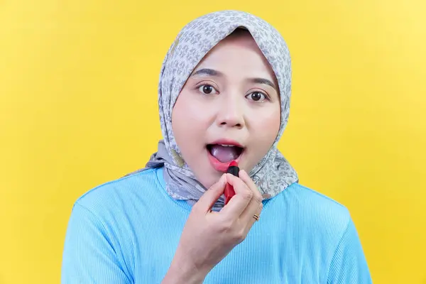 Tesettürlü Asyalı bir kadın kameraya bakarken kırmızı ruj sürüyor, sarı arka planda mavi bir kıyafet giyiyor..