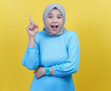 Tesettür ve mavi gömlekli neşeli Asyalı bir kadın işaret parmağını kaldırarak heyecan ve merakını dile getiriyor..