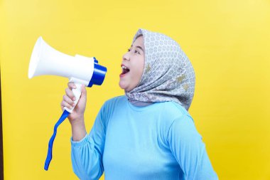 Mavi ve tesettürlü Asyalı bir kadın elinde bir megafonla sarı arka planda coşkuyla konuşuyor..