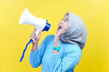 Mavi elbiseli neşeli bir Asyalı kadın elinde bir megafonla sarı arka planda güvenle konuşuyor..