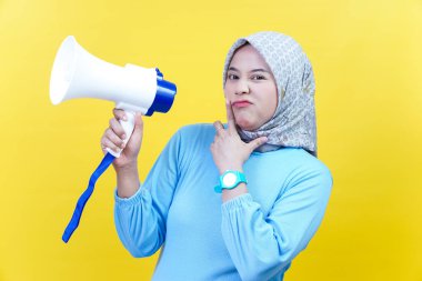 Tesettürlü ve mavi elbiseli Asyalı bir kadın megafonu tutarken düşünceli ve neşeli bir ifade takınıyor..