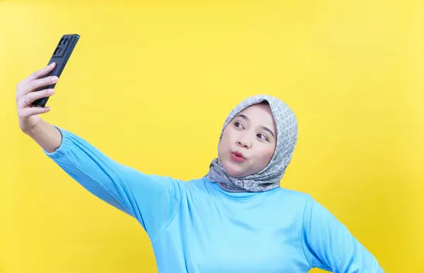 Tesettürlü ve mavi tişörtlü neşeli Asyalı bir kadın selfie çeker, gülümser ve telefonuna bakar..